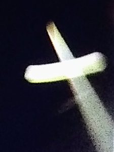 A wide night photograph capturing a massive, prominent cross shape made of intense, radiant white light shining brightly in the open sky.