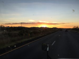 Silhouetted travelers moving toward a distant mountain range beneath a vibrant orange and yellow sunset.