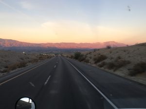 : A striking view looking down an open road on a highway at dusk, with an orange-gold sunset on the horizon and mountains in the far distance under a framing sky.