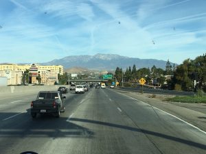 A striking photograph taken from a vehicle driving down a busy highway toward a big city skyline with trees lining the road and towering mountains far off in the background.