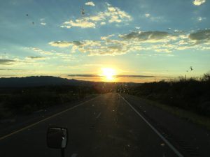 Open highway extending toward a vibrant orange and gold sunset on the horizon with distant mountain silhouettes under dark twilight clouds.
