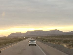 An open highway drive at dusk with a car ahead under heavy dark clouds and a glowing sunset at the bottom of the horizon.