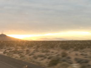 A moving-vehicle highway shot capturing a vivid peach and gold sunset sky stretching out over sweeping mountain peaks in the far distance on the driver's right side.