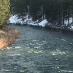 A beautiful river perspective looking out from the bank at moderate rapids, with a dense forest of evergreen trees and fresh snow covering the opposite side of the water.