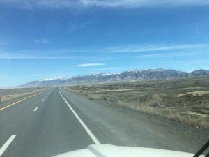A wide landscape photograph looking down a clear highway with two lanes moving forward and two lanes on the opposite side, beneath a beautiful mountain range stretching all the way across the horizon under a soft sky.