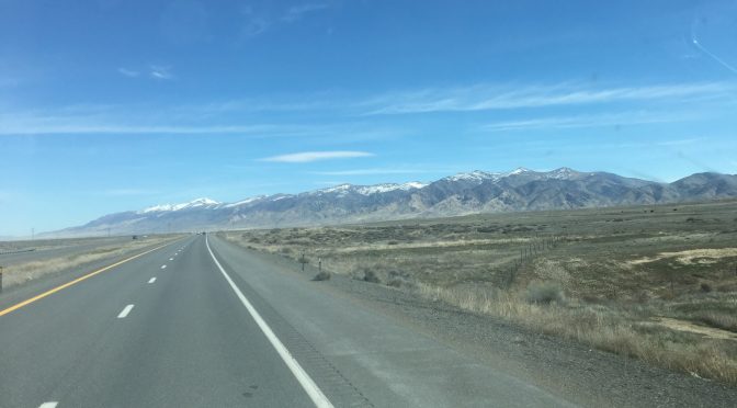 A wide landscape photograph looking down a clear highway with two lanes moving forward and two lanes on the opposite side, beneath a beautiful mountain range stretching all the way across the horizon under a soft sky.