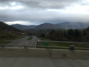  A road-trip photograph taken from a highway overpass, looking out to the right at a majestic mountain range standing tall in the far background under a twilight sunset.