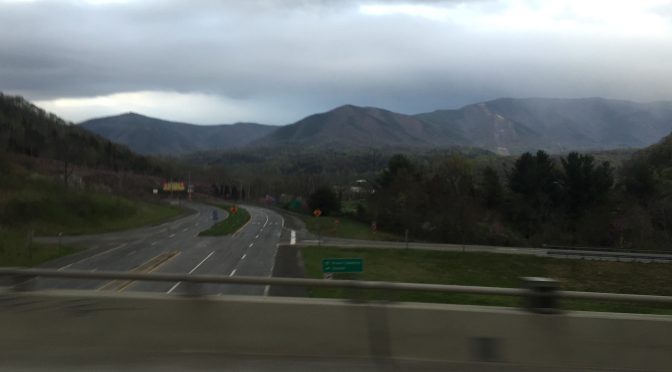 A road-trip photograph taken from a highway overpass, looking out to the right at a majestic mountain range standing tall in the far background under a twilight sunset.