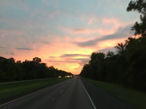 A spectacular landscape photograph taken looking down a divided highway with two lanes moving forward and two oncoming lanes, perfectly framed by tall trees on both sides under a vibrant evening sunset sky.
