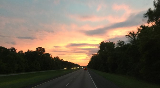 A spectacular landscape photograph taken looking down a divided highway with two lanes moving forward and two oncoming lanes, perfectly framed by tall trees on both sides under a vibrant evening sunset sky.