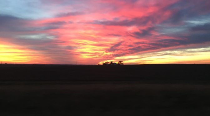 A beautiful dusk sunset showing a vast sky filled with gold, pink, and light blue clouds casting a soft glow over a quiet landscape.
