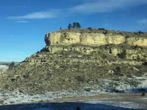  A landscape photograph from a traveler's journey showing a massive solid rock mountain rising behind an open field with a few trees clustered at the very top.