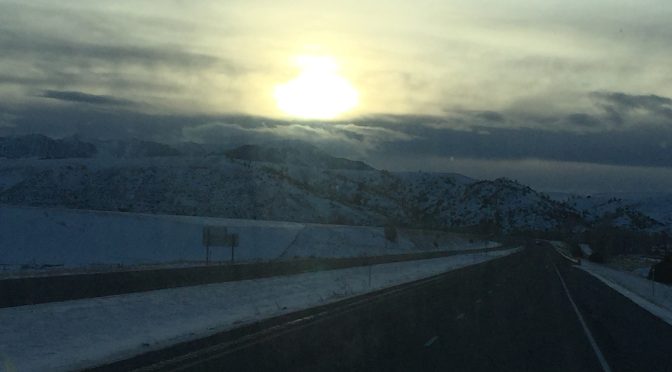 A high-quality, inspiring landscape photograph of a bright, radiant sun peaking over the dark silhouette of a rugged mountain range at dawn, casting warm golden rays across a dark sky.