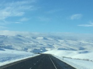 A high-quality landscape photograph of a clear road heavily lined with crisp white snow, leading toward a towering, snow-covered mountain range under a vast sky.