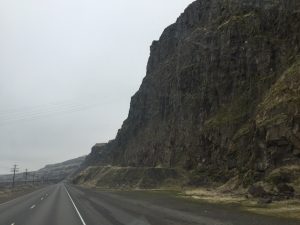 A dramatic landscape perspective looking down an asphalt road with a massive, towering sheer rock mountain rising steeply on the right side