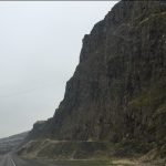 A dramatic landscape perspective looking down an asphalt road with a massive, towering sheer rock mountain rising steeply on the right side.