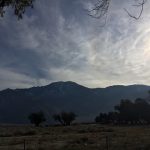 A beautiful photograph of a wide open field with majestic mountains in the background, a few peaceful trees, and a big sun shining in the right corner under a dark sky.