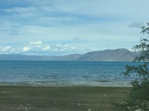 A stunning, expansive view of a deep blue lake. Across the wide water stands a grand, majestic mountain, creating a perfect balance of unmovable strength and peaceful reflection under a bright sky.