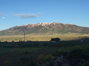A beautiful country ranch featuring a vibrant green field in the foreground, stretching toward a majestic range of snow-capped mountains in the far distance with a large, beautiful moon shining on the left-hand side.
