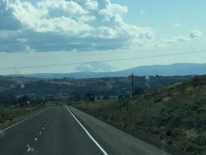 A striking perspective looking down an open highway toward a majestic mountain range rising in the distance, with a massive rock mountain visible in the center under a clear sky.
