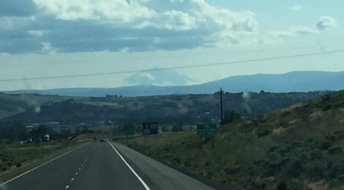 A striking perspective looking down an open highway toward a majestic mountain range rising in the distance, with a massive rock mountain visible in the center under a clear sky.