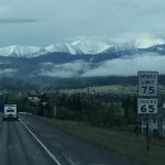 A beautiful view looking down a road heading into a peaceful town, with majestic rock mountains standing tall in the front and soft white clouds framing the peaks above.