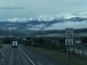 A beautiful view looking down a road heading into a peaceful town, with majestic rock mountains standing tall in the front and soft white clouds framing the peaks above.