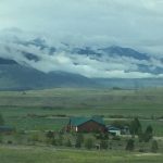 A breathtaking view across a vast field where a peaceful farmhouse rests in the middle, with an immense rock mountain range and low clouds stretching all the way across the horizon behind it.