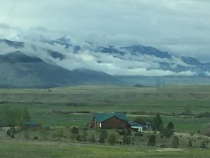 A breathtaking view across a vast field where a peaceful farmhouse rests in the middle, with an immense rock mountain range and low clouds stretching all the way across the horizon behind it.