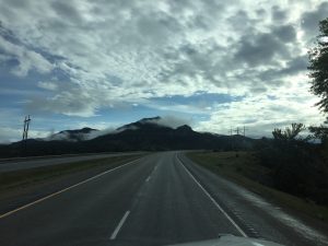 A view from inside a vehicle driving along a high-elevation highway curve, looking toward two massive mountain peaks rising directly ahead and partially hidden by low clouds.