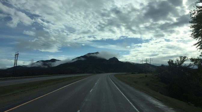 A view from inside a vehicle driving along a high-elevation highway curve, looking toward two mountain peaks rising directly ahead and partially hidden by low clouds.