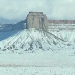 A beautiful photograph of a massive, solid rock mountain with snow on its peaks under a vast sky, with no pavement or roads visible.