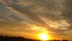 A wide view of expansive flat desert land beneath a fire-hot sky filled with dramatic clouds, dominated by a massive, intensely bright yellow and gold sun.