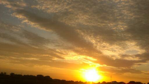A wide view of expansive flat desert land beneath a fire-hot sky filled with dramatic clouds, dominated by a massive, intensely bright yellow and gold sun.