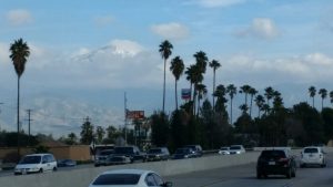 A peaceful town road with cars parked on both sides, leading toward a view of massive snow-capped mountains rising gracefully in the far left distance under a clear sky.