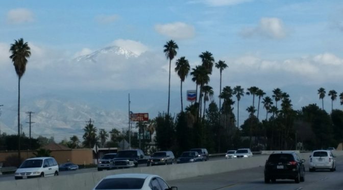 A peaceful town road with cars parked on both sides, leading toward a view of massive snow-capped mountains rising gracefully in the far left distance under a clear sky.
