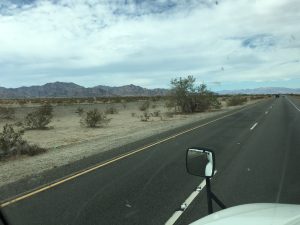 A view from inside a moving car, looking through the front windshield on a gray, overcast day. A prominent ridge of mountains stretches out completely along the left side of the frame, reaching all the way forward toward the distance under a heavy blanket of low clouds.