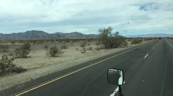 View from inside a vehicle driving down a highway with a large mountain range spanning the entire left side under an overcast, cloudy sky.