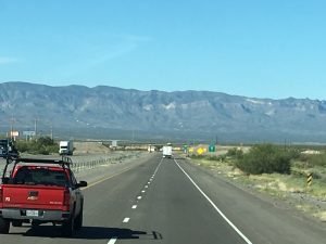 A long, open stretch of empty highway cutting forward through a vast landscape with a large mountain range stretching across the horizon miles away.