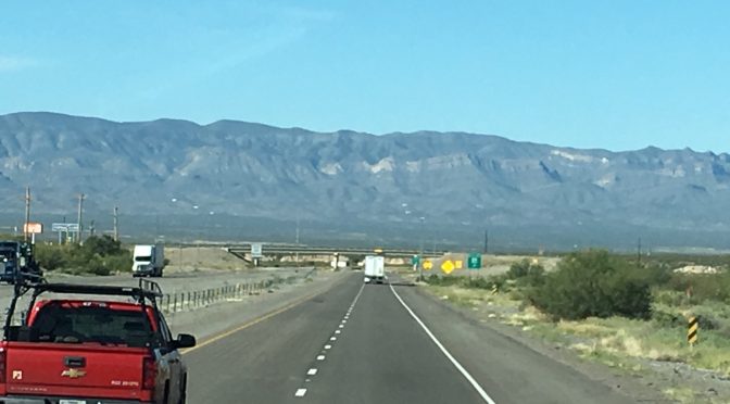 A long, open stretch of empty highway cutting forward through a vast landscape with a large mountain range stretching across the horizon miles away.