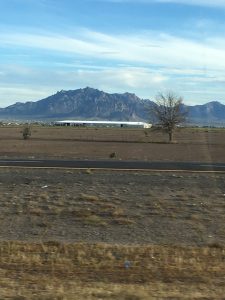 A beautiful wide-open desert field with sparse green brush leading to a majestic, rugged rock mountain range stretching across the horizon under an overcast sky.