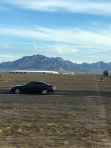 •  A dramatic, wide-perspective road photograph taken from a moving vehicle showing a black car driving past a large industrial warehouse in the foreground, with massive, rugged mountain peaks rising sharply against the horizon in the far distance.