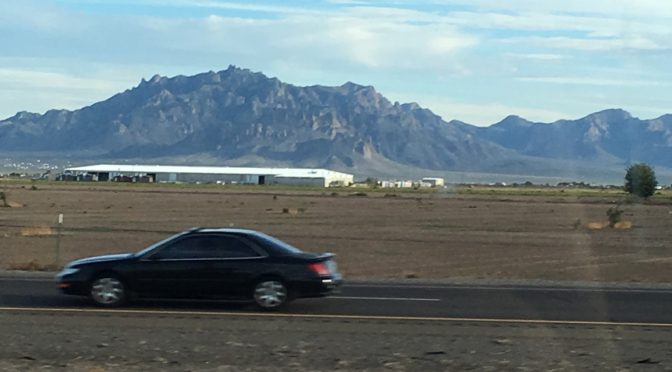 A dramatic, wide-perspective road photograph taken from a moving vehicle showing a black car driving past a large industrial warehouse in the foreground, with massive, rugged mountain peaks rising sharply against the horizon in the far distance.