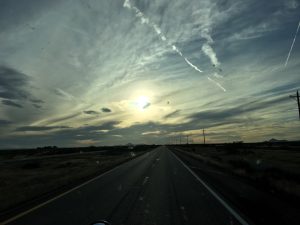 A beautiful dusk sunset showing an open asphalt road stretching forward to a majestic rock mountain range rising silhouette against a gold, pink, and light blue sky in the far distant horizon.
