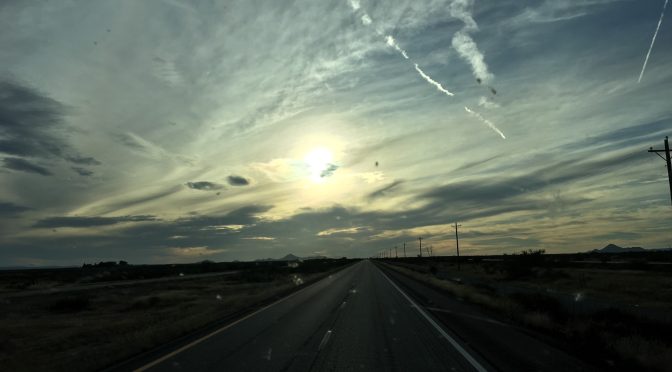 A beautiful dusk sunset showing an open asphalt road stretching forward to a majestic rock mountain range rising silhouette against a gold, pink, and light blue sky in the far distant horizon.