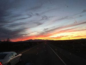 A panoramic desert landscape at sunset featuring soft, swirly clouds swept across one side of the sky in a mixture of bright orange, radiant yellow, and warm peach tones, with a rugged mountain range standing silhouette miles away in the distance.