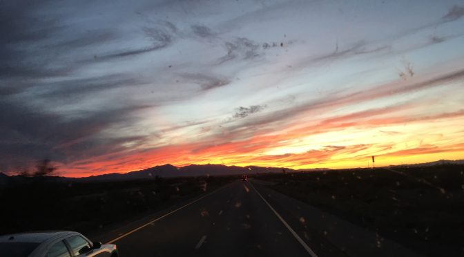 A panoramic desert landscape at sunset featuring soft, swirly clouds swept across one side of the sky in a mixture of bright orange, radiant yellow, and warm peach tones, with a rugged mountain range standing silhouette miles away in the distance.
