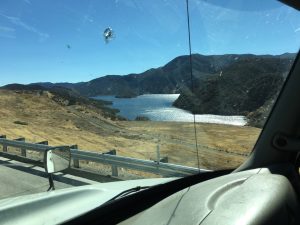 A view looking out the side window from a moving vehicle down an open asphalt highway beside a passing body of water, with a rugged mountain range standing tall in the far background under a clear sky.
