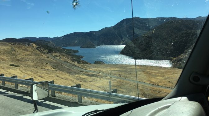 A view looking out the side window from a moving vehicle down an open asphalt highway beside a passing body of water, with a rugged mountain range standing tall in the far background under a clear sky.