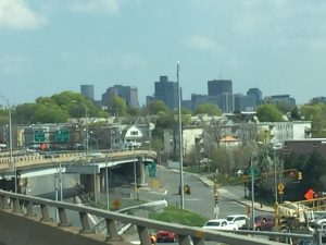 An atmospheric view from an expressway with a city skyline, street layouts, and tall buildings visible in the background